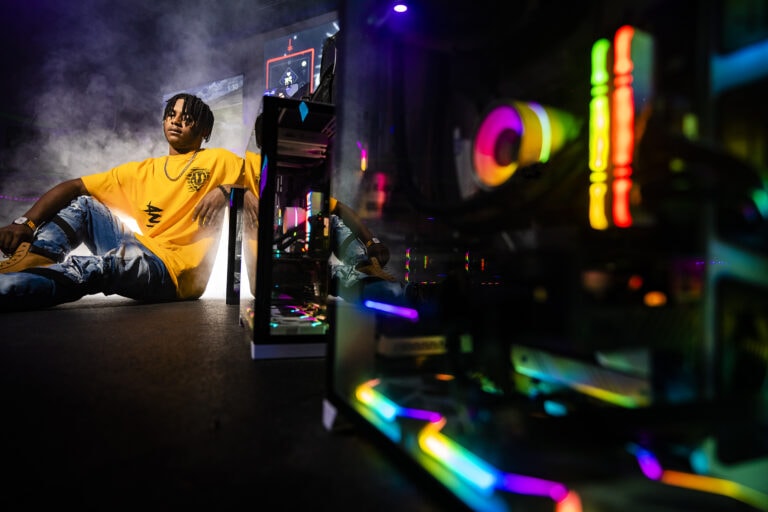 Senior guy leans against a row of custom built gaming PCs at the Vyral Compound for his gamer senior photos in Kettering, Ohio.