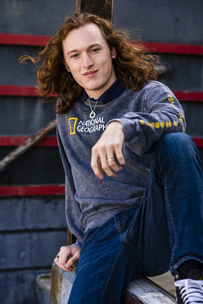 Senior guy from Carroll High School wears a National Geographic sweater and sits on a stoop in downtown Dayton during his senior pictures.