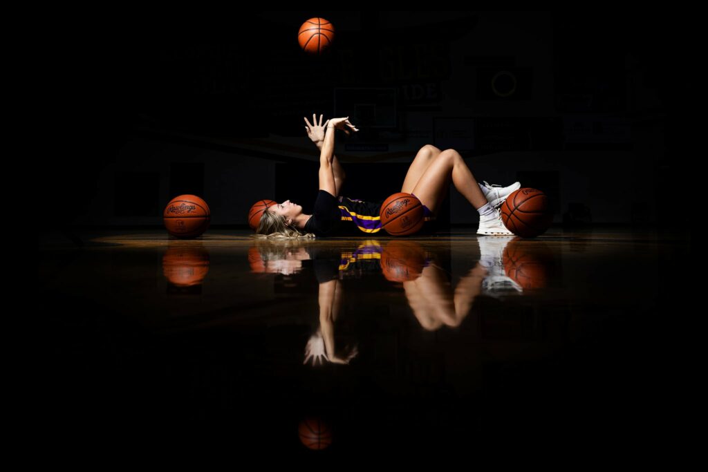Senior high school basketball player, Taylor Scohy, is lying on the Bellbrook High School basketball court with several basketballs around her. She is shooting a basketball into the air while wearing her black uniform.