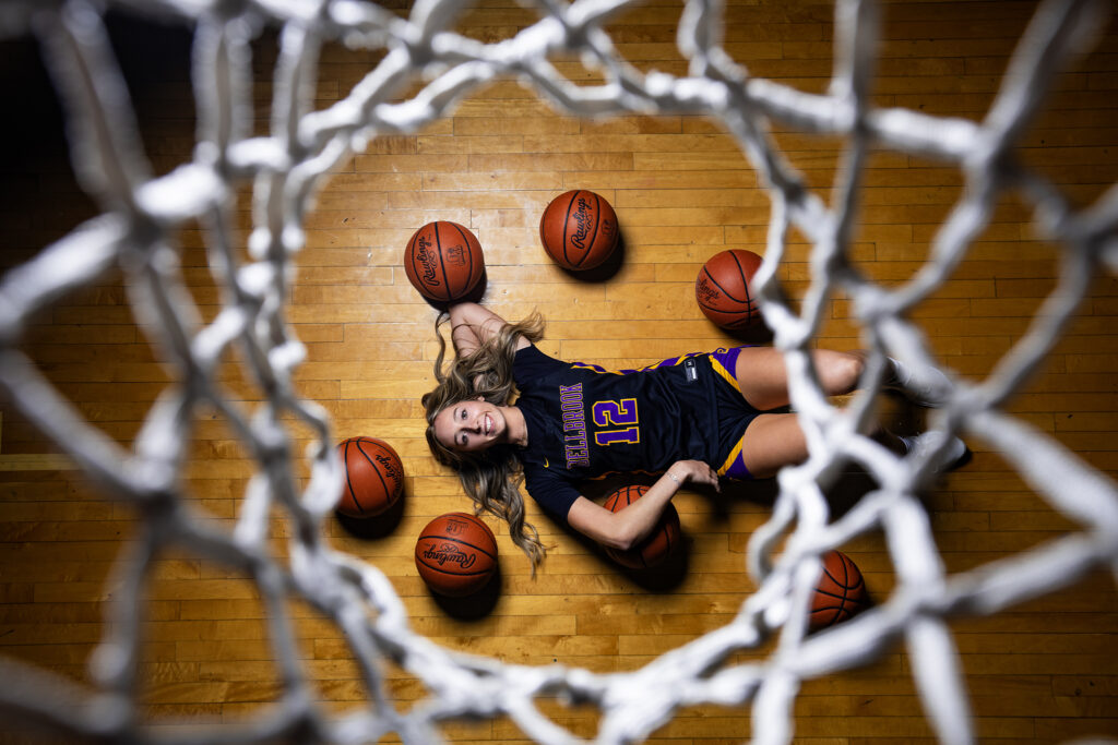 Bellbrook High School's Taylor Scohy lays on the basketball court surrounded by basketballs, wearing her black uniform. The photo is taken from above, looking down on her through a basketball hoop net.