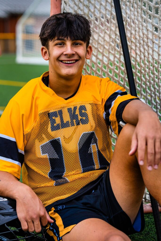 Centerville High School senior lacrosse player Seth Alejandrino sits against a lacrosse goal smiling. He is wearing a yellow and black elks lacrosse uniform.