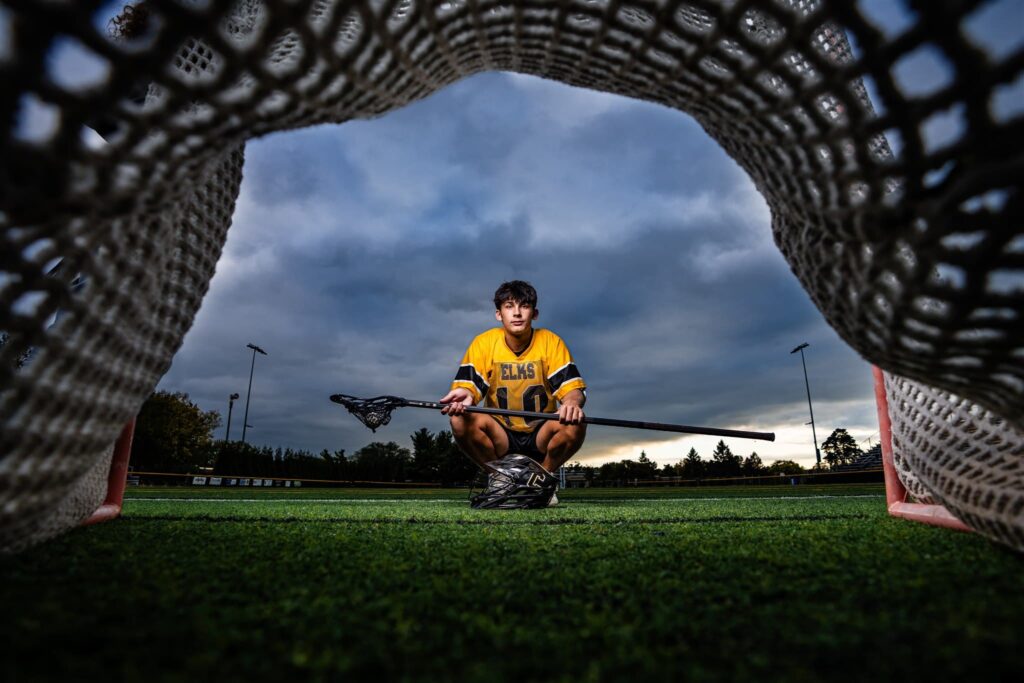 Seth Alejandrino, a senior at Centerville High School, squats in front of a lacrosse goal holding a defensive long pole for his senior pictures. He is wearing a black and gold lacrosse uniform, and his lacrosse helmet is sitting on the turf in front of him.