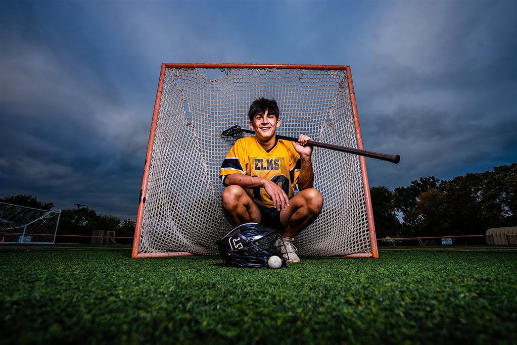 Centerville High School senior lacrosse player Seth Alejandrino squats in front of a lacrosse goal at Alumni Stadium on a stormy evening. He is wearing a gold and black uniform and holding his defensive long pole over his shoulder while smiling.
