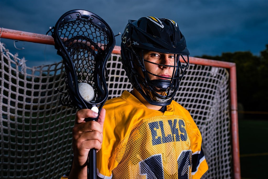 Centerville High School senior Seth Alejandrino stands in front of a lacrosse goal in full uniform, looking stoically at the camera. He is wearing a gold and black uniform and a black helmet, with a lacrosse defensive long pole in his right hand. There is a white lacrosse ball in the pocket of the stick.