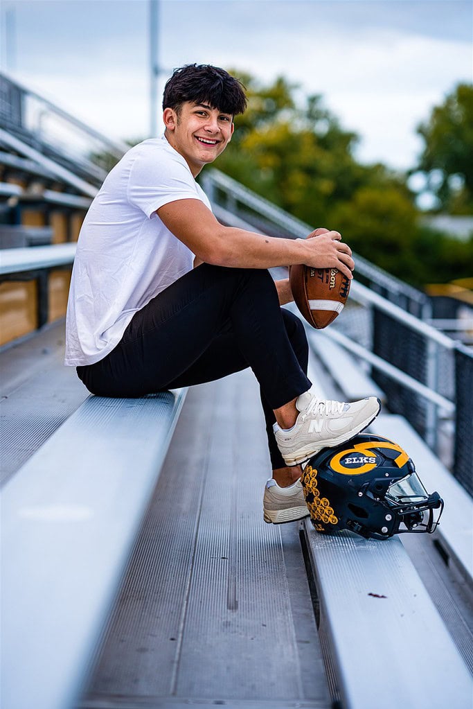 Seth Alejandrino, a two-sport senior athlete at Centerville High School, sits in the stadium bleachers at Alumni Stadium for senior pictures. He is wearing a white shirt and black joggers with white sneakers, with one foot on his black football helmet. He smiling at the camera while holding a football.
