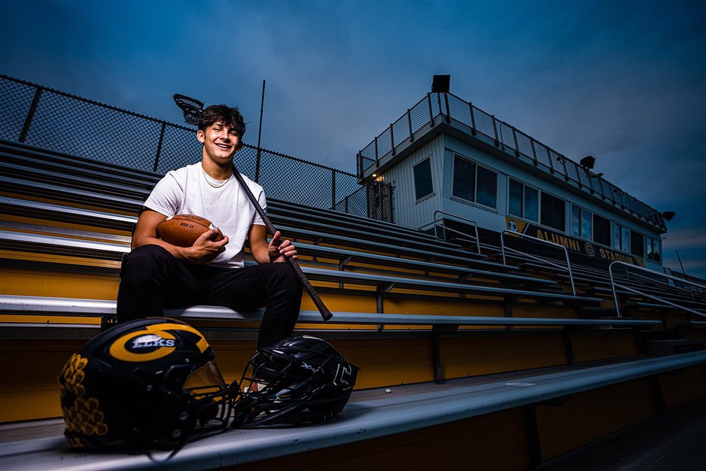 Two sport senior athlete Seth Alejandrino sits in the stands at Alumni Stadium during his senior photos. He is wearing a white t-shirt and black joggers. He is holding a football in his right arm, and his defensive long pole for lacrosse in his left. Both his football and lacrosse helmets are sitting in front of him.