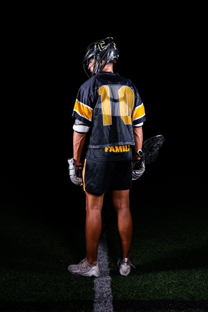 Seth Alejandrino stands with his back to the camera, looking over his shoulder at nighttime during his senior photos. He is wearing a black with gold accents lacrosse uniform.