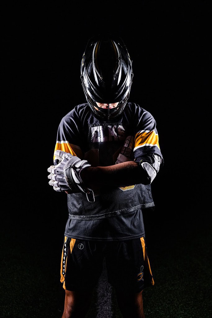 Seth Alejandrino looks down in his full black Centerville Elks lacrosse uniform. He is lit from the sides, and it is black all around him.