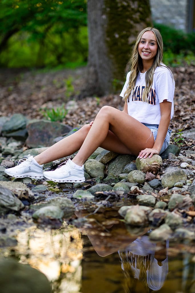 Taylor Scohy, a senior athlete at Bellbrook High School, sits by a rocky stream for her senior photos with Rev Studios. She is wearing jean shorts, white sneakers, and a white UT-Martin t-shirt.