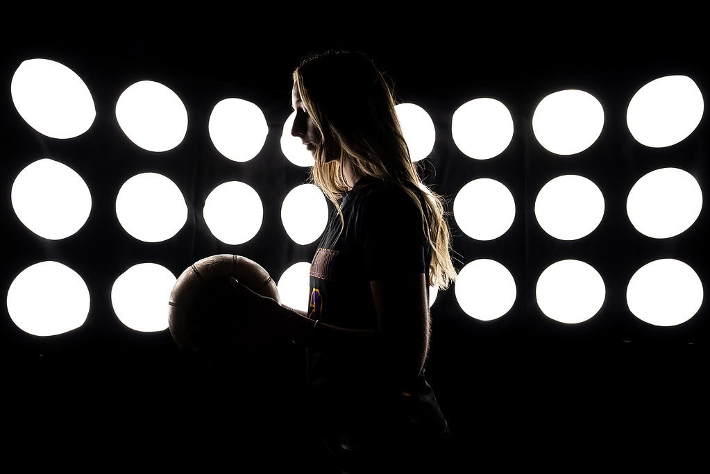 Silhouetted against stadium lights, Bellbrook High School Senior Basketball Player Taylor Scohy stands in profile, looking down at a basketball.