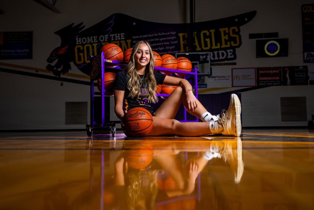 Bellbrook High School basketball player Taylor Scohy sits on the court, leaning against a basketball rack, wearing her all-black uniform