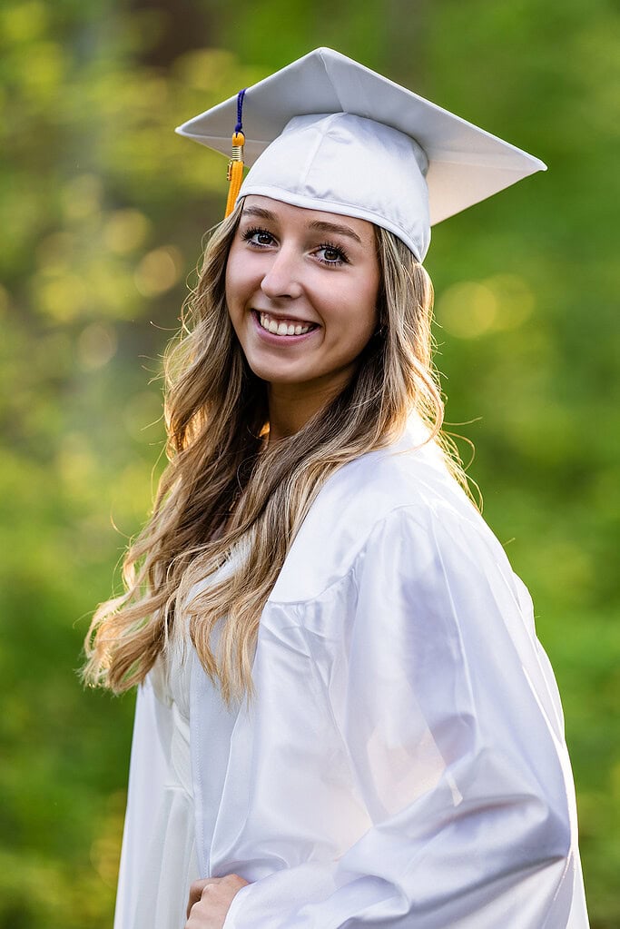 Taylor Scohy, a senior at Bellbrook High School, stands proudly smiling in a white graduation cap and gown outdoors in warm sunlight.